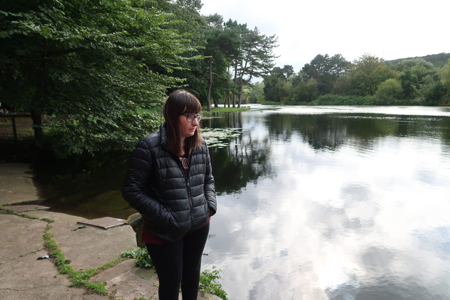 Holly stood at the edge of a lake with her hands in her pockets, she is wearing a black padded jacket and black leggings and is looking towards the lake