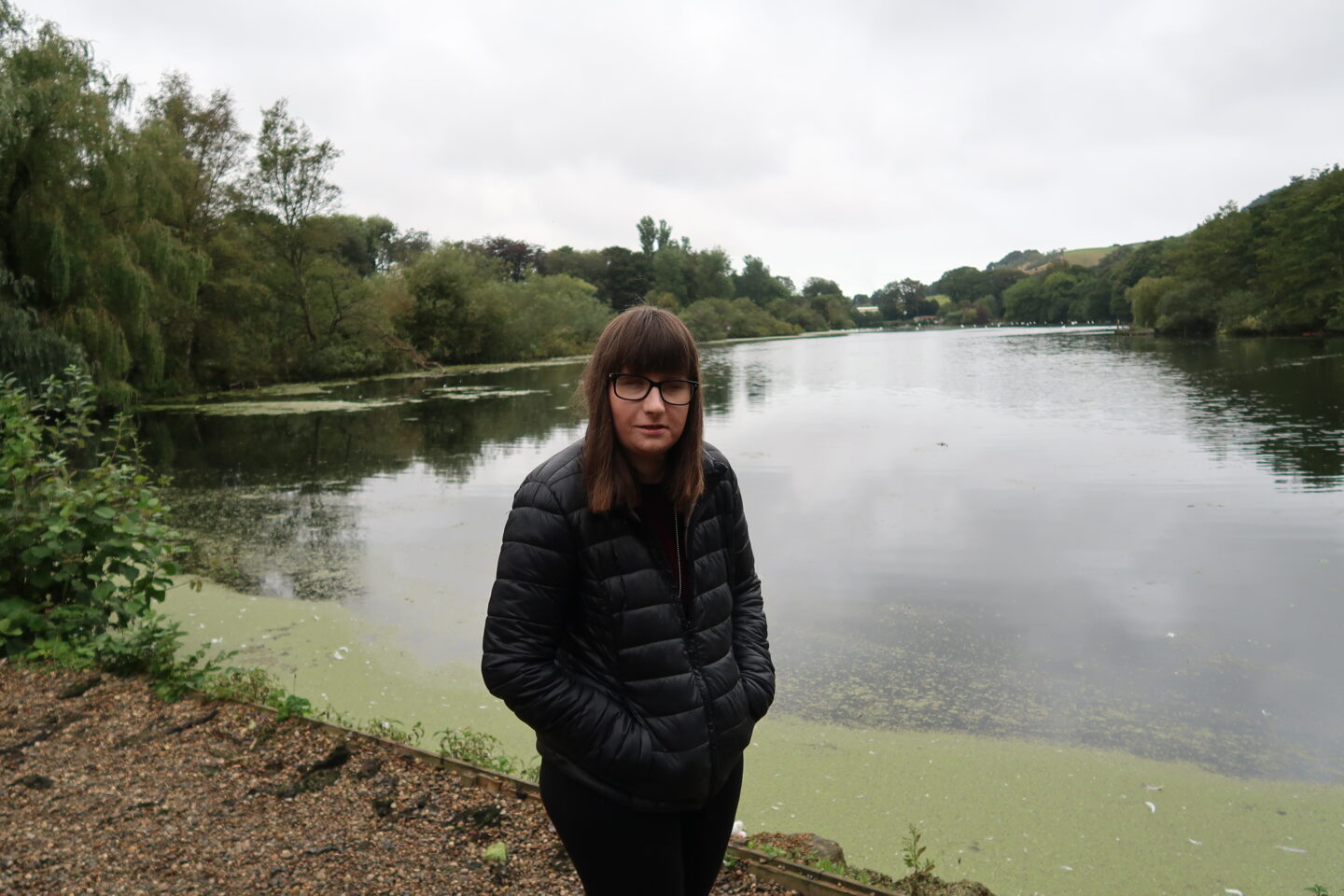 Holly stood on a gravel path with her hands in her pockets and is smiling. She is wearing a black padded jacket, black leggings and trainers. There is a lake and a mass of lovely green trees in the background