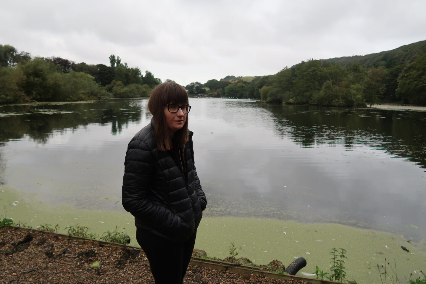 Holly stood on a gravel path, she has her hands in her pockets and is looking to the side, there is a lake and green trees in the distance. She is wearing a black jacket, black leggings and trainers