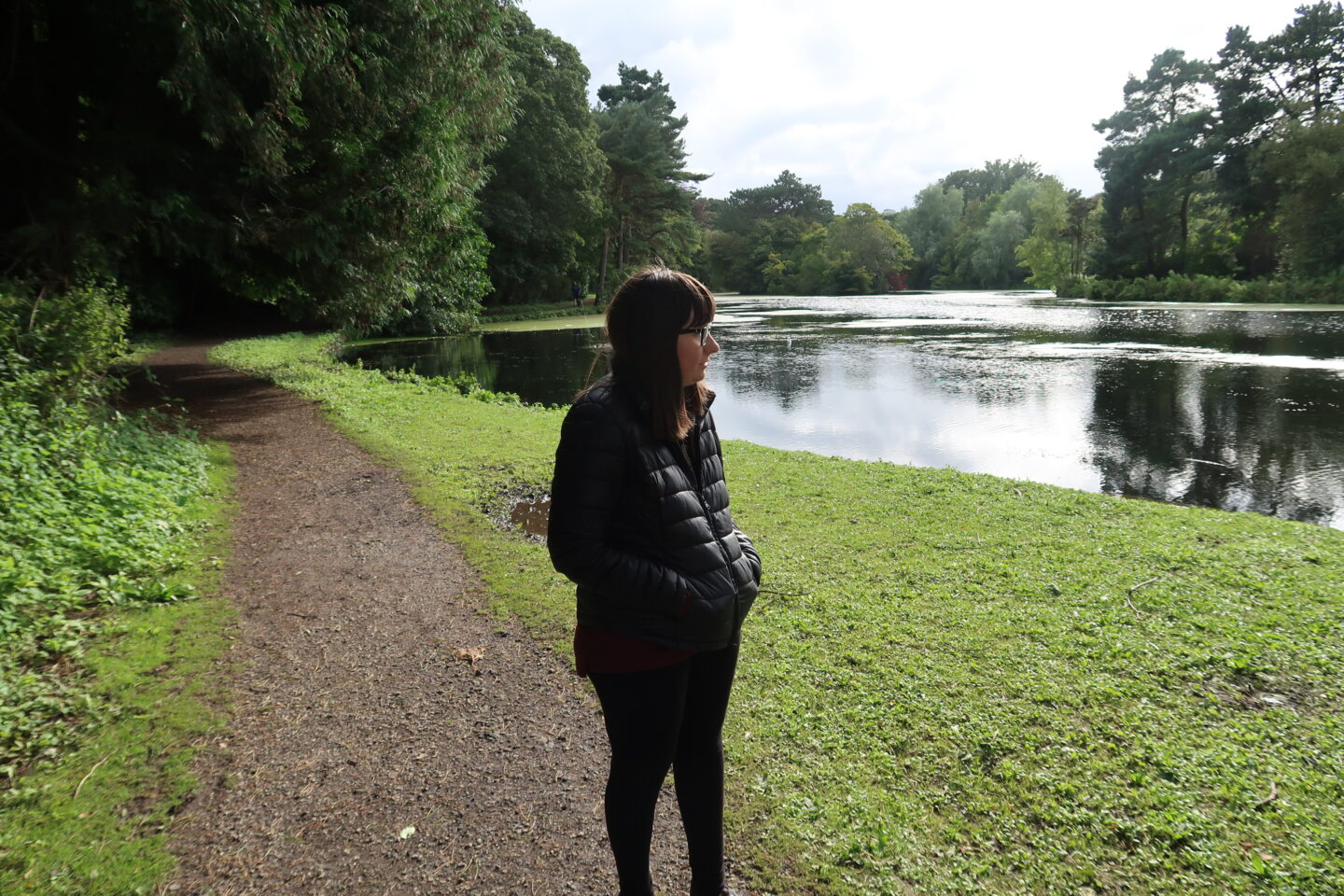 Holly stood on a gravel path with her hands in her pockets and looking towards a stunning lake. She is wearing a black padded jacket and black leggings.