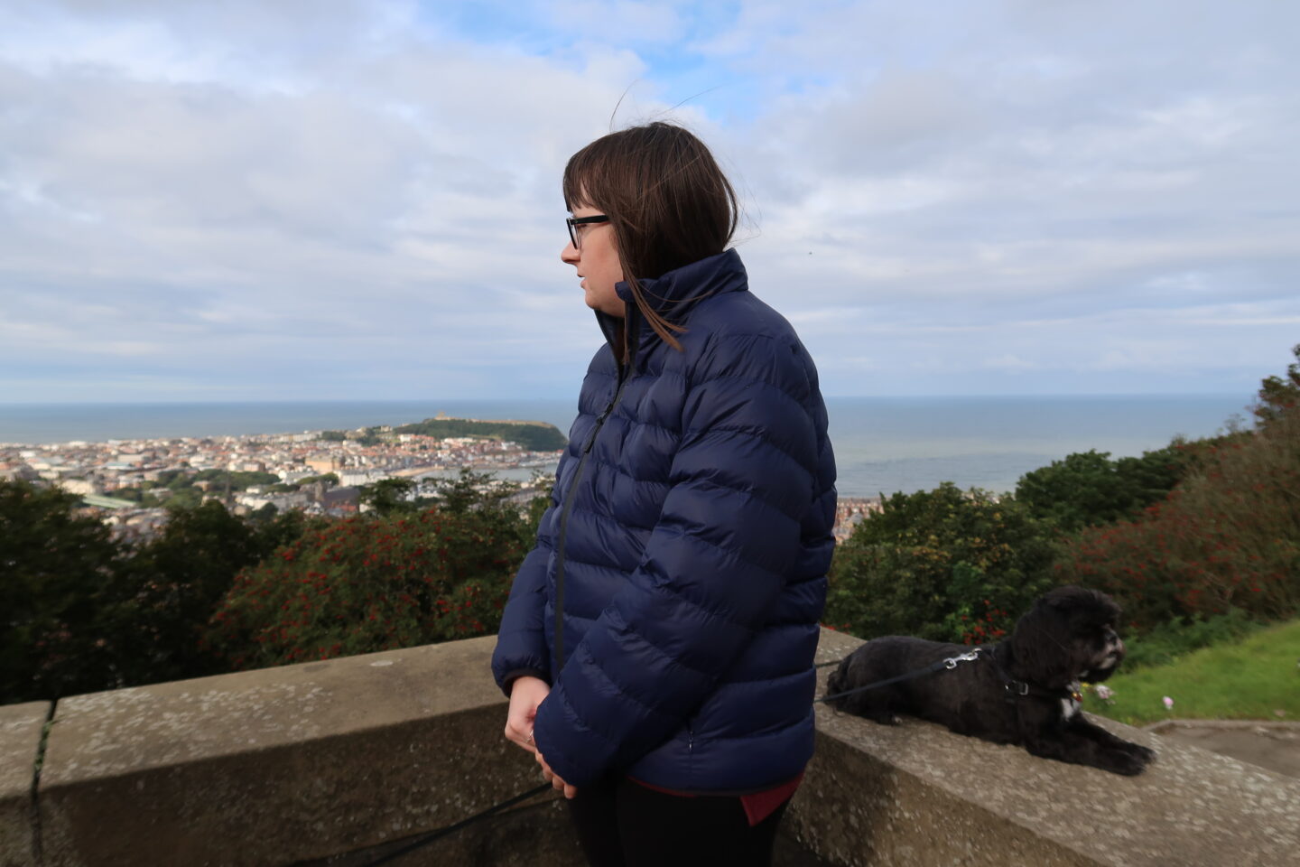 Holly leant against a stone wall with her hands crossed in front of her, she is wearing a blue padded jacket and black leggings. Her small black dog is on the wall behind her. The sea, a bay and trees can be seen in the distance