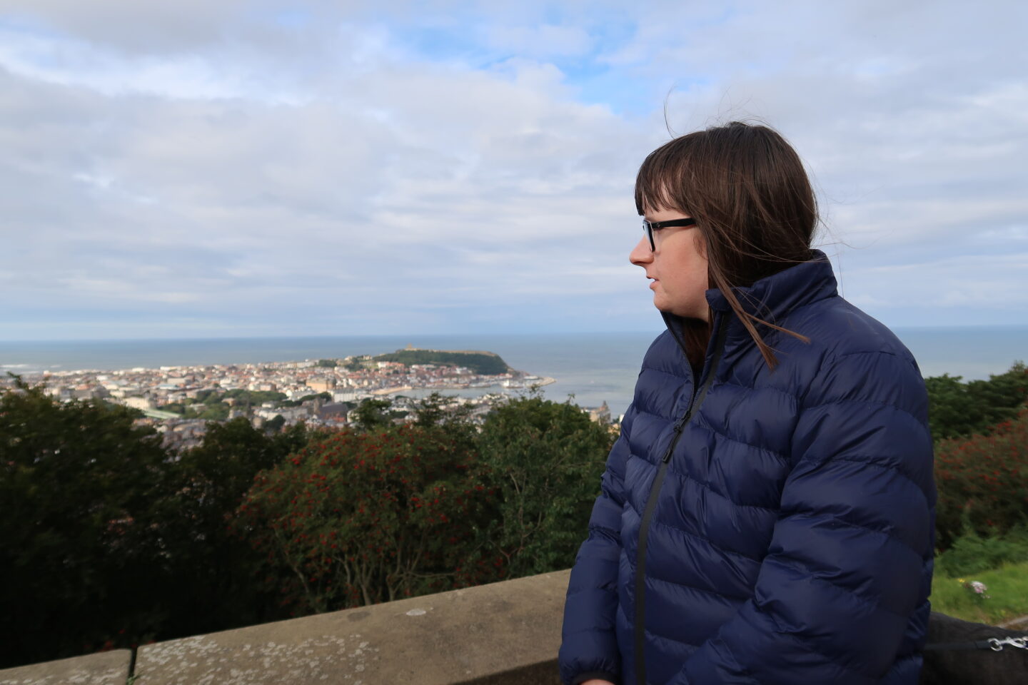 Holly leant against a stone wall looking to the side, she is wearing a blue padded jacket. A mass of trees and a bay can be seen in the distance