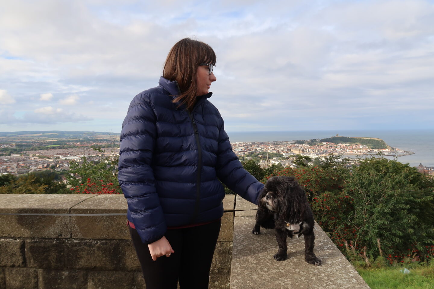 Holly stood against a stone wall with her hand on her small black dog, she is wearing black leggings and a blue padded jacket. There is a bay and a mass of green trees in the distance