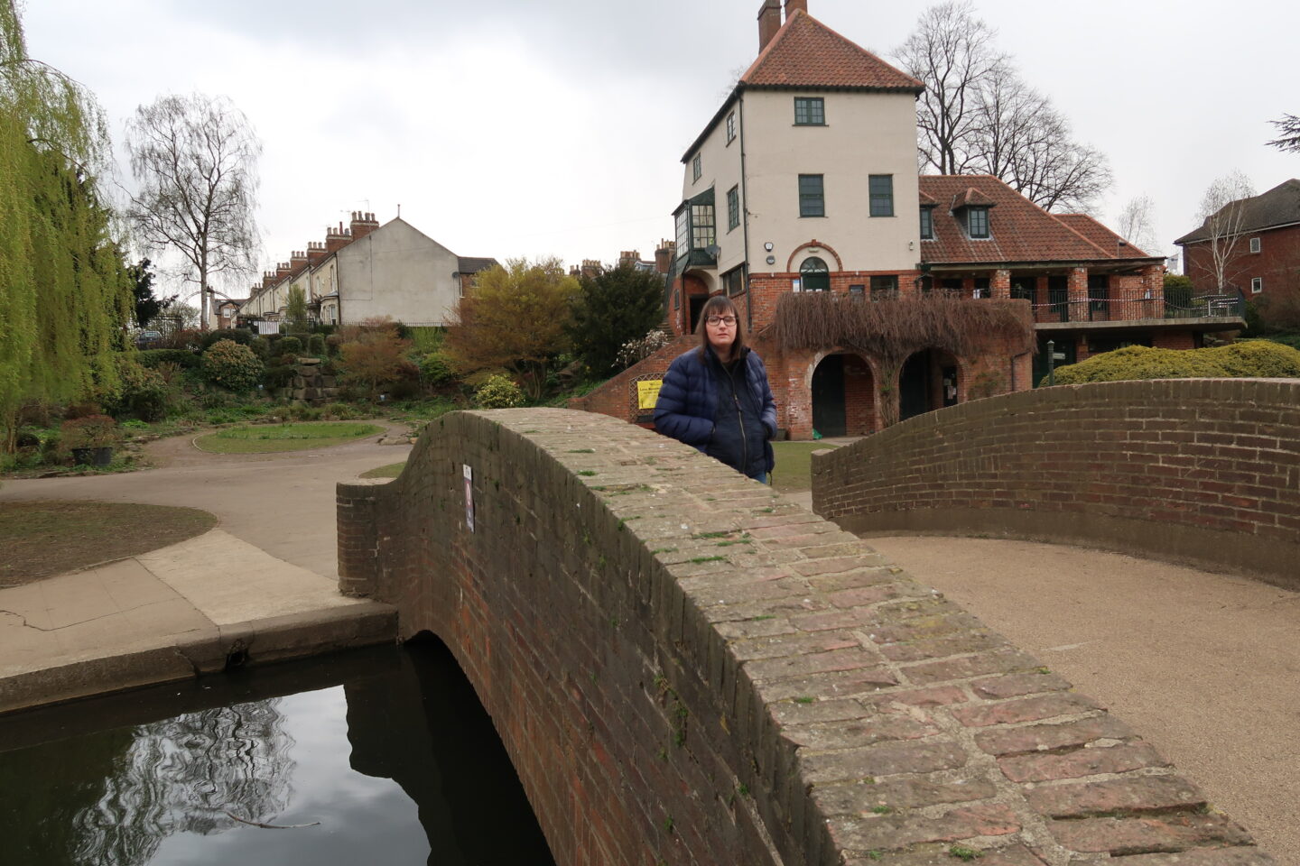 Holly stood on a stone bridge with her hands in her pockets, she is wearing a blue padded jacket and blue jeans. There is a stone building in the background