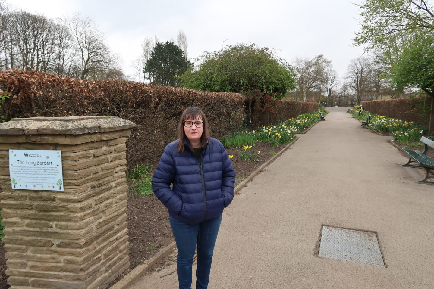 Holly stood on a stone path with daffodils and brown hedges at either side, she is wearing a blue padded jacket, blue jeans and grey trainers