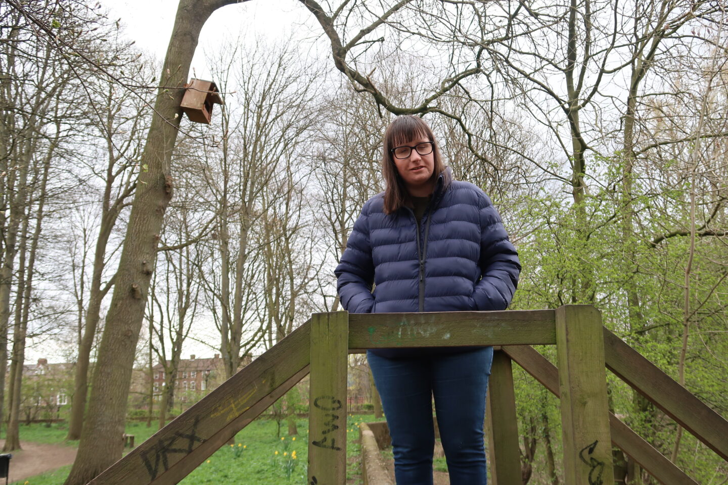 Holly stood on a wooden bridge with her hands in her pockets and looking down, she is wearing a blue padded jacket and blue jeans. There is a mass of green trees in the background, clouds can be seen peering through the trees