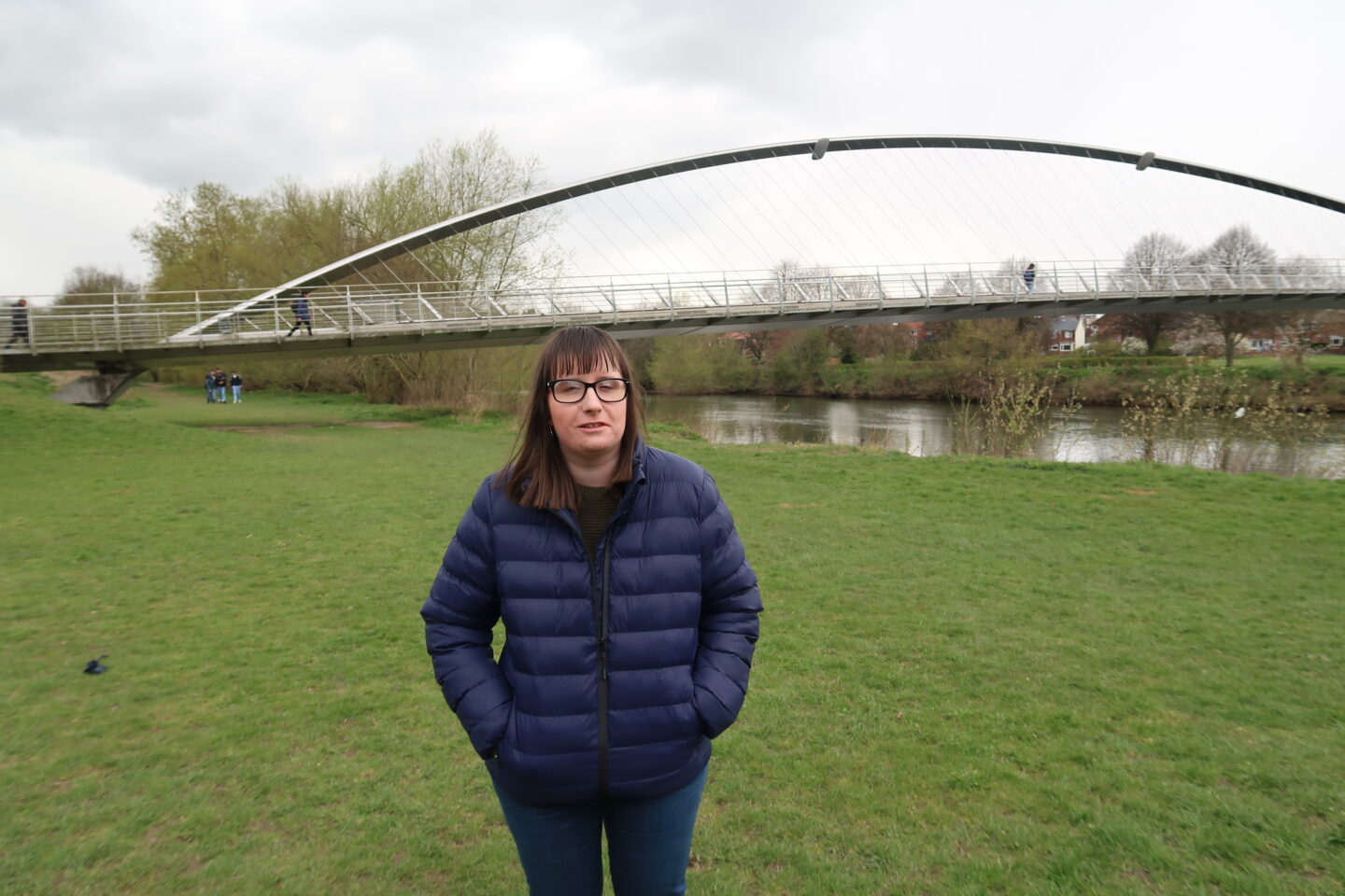 Holly stood on some grass with her hands in her pockets, she is wearing a blue coat and blue jeans. There is a river and trees in the background and a tall metal bridge in the distance