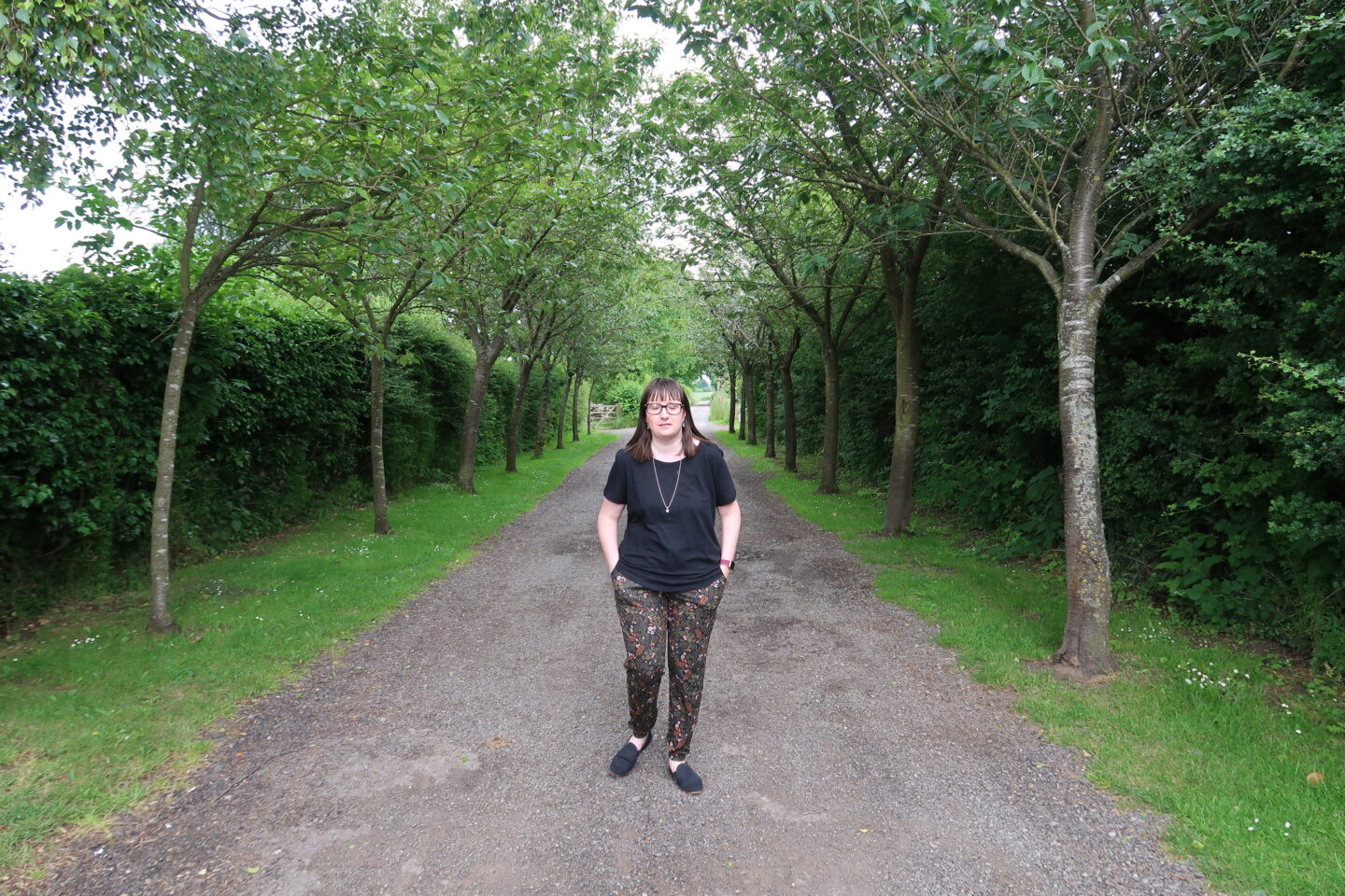 Holly stood in a tunnel of trees. She's wearing a black top and multicoloured trousers and has her hands in her pockets