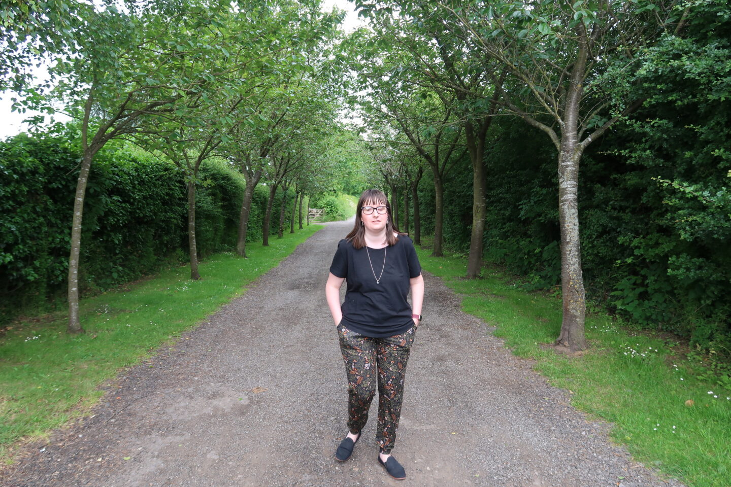 A close-up photo of Holly stood in a tunnel of trees with her hands in her pockets, she is wearing a black top and multicoloured trousers