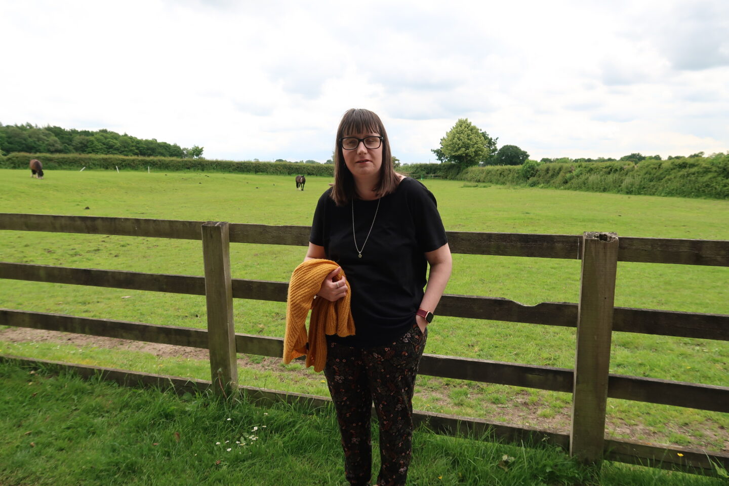 Holly stood in front of a fence, a green field can be seen in the distance. She is wearing a black top and multi-coloured trousers and is holding a yellow cardigan over her right arm
