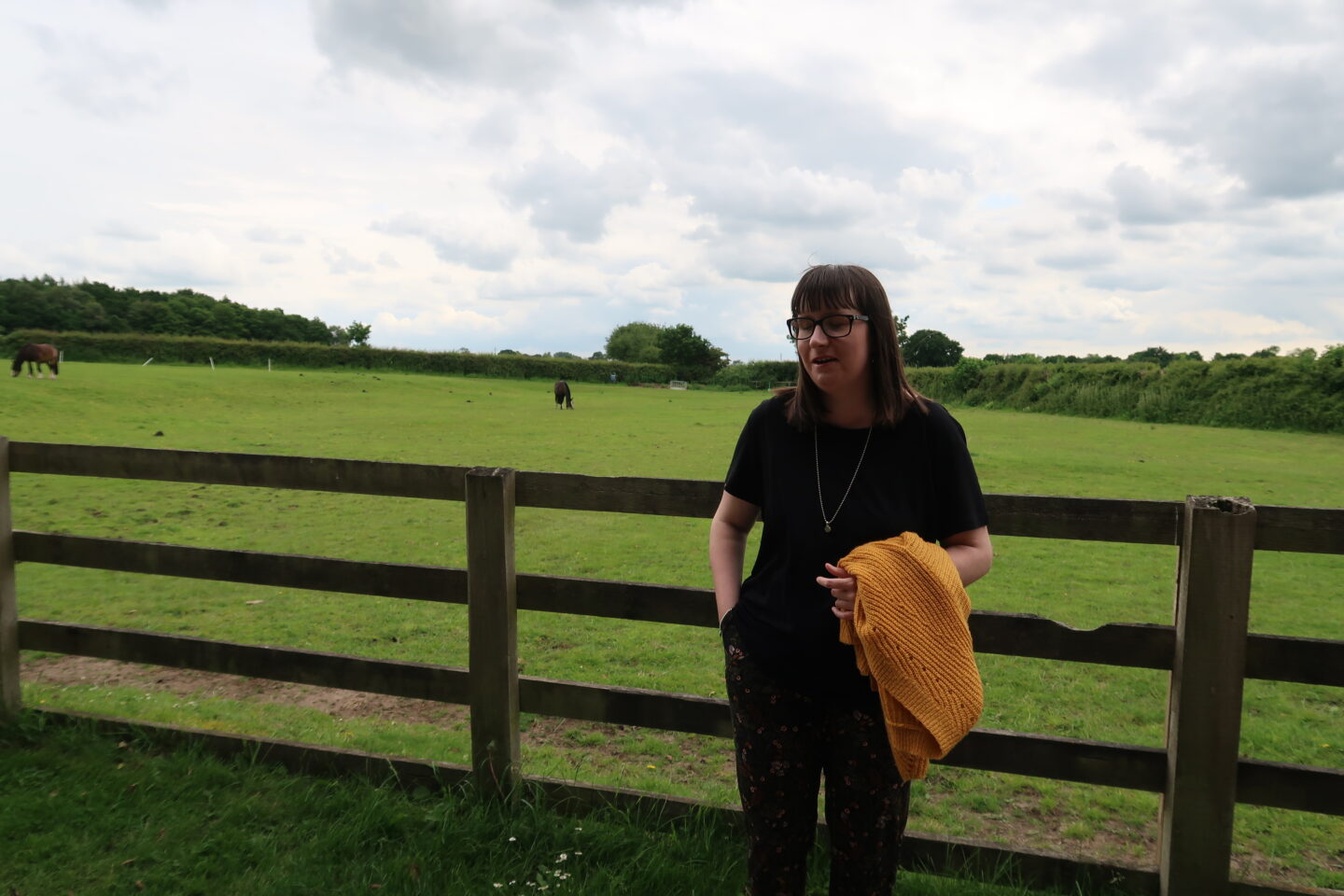 Holly stood in front of a fence, there is a green field in the distance. She's wearing a black top and multi-coloured trousers. She is holding a yellow cardigan over her left arm and is looking slightly to the right