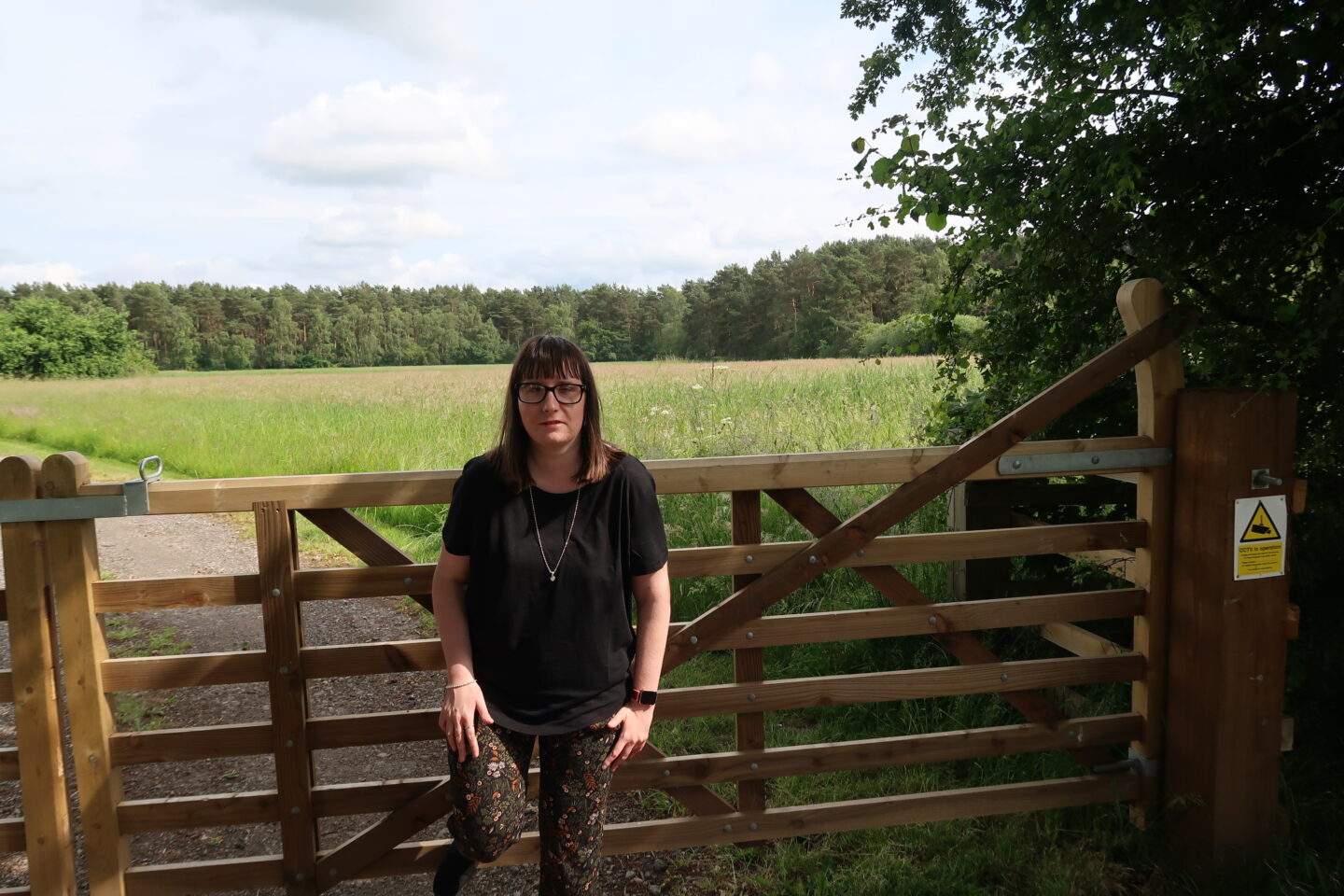 Holly stood with her foot up on a gate, she is wearing a black top and multi-coloured trousers. There is a green field and woodland in the distance