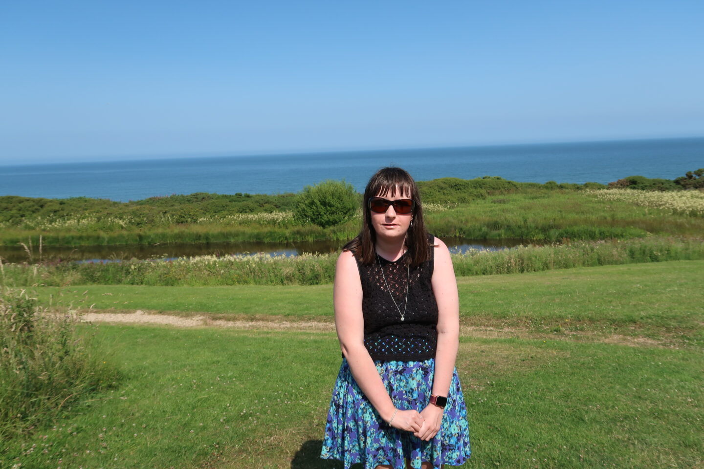 A close-up photo of Holly stood with her hands clasped together in front of her, she is wearing a blue skirt, a black crocheted top, a necklace and sunglasses. There is a grassy area and a lake behind her, the sea can be seen in the distance
