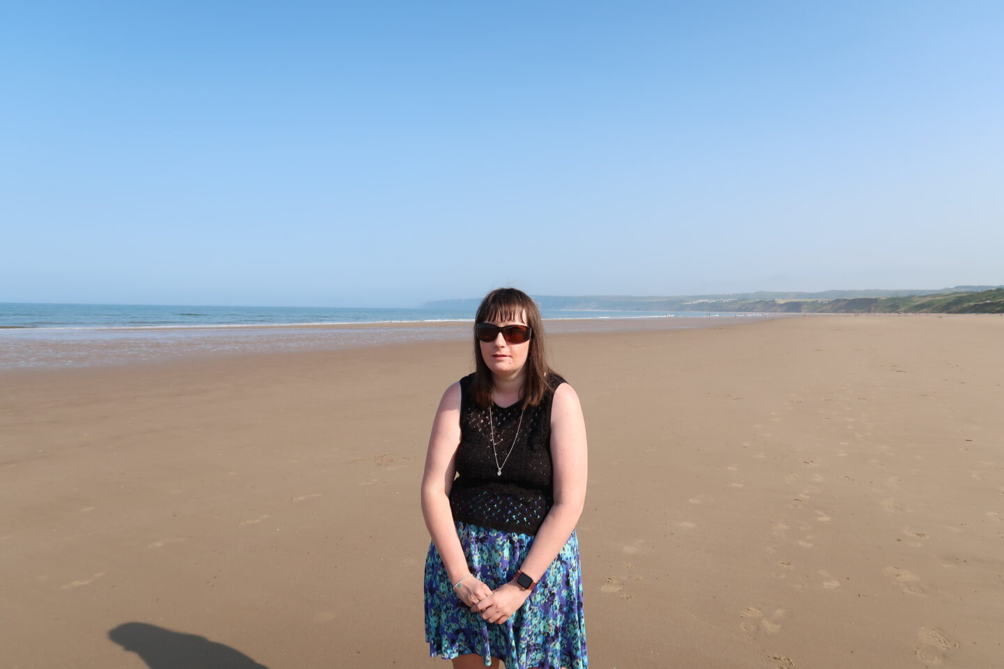 A close-up photo of Holly stood on a beach, the sea is in the background. She has her hands clasped in front of her and is wearing a blue skirt, a black crocheted top, a necklace and sunglasses