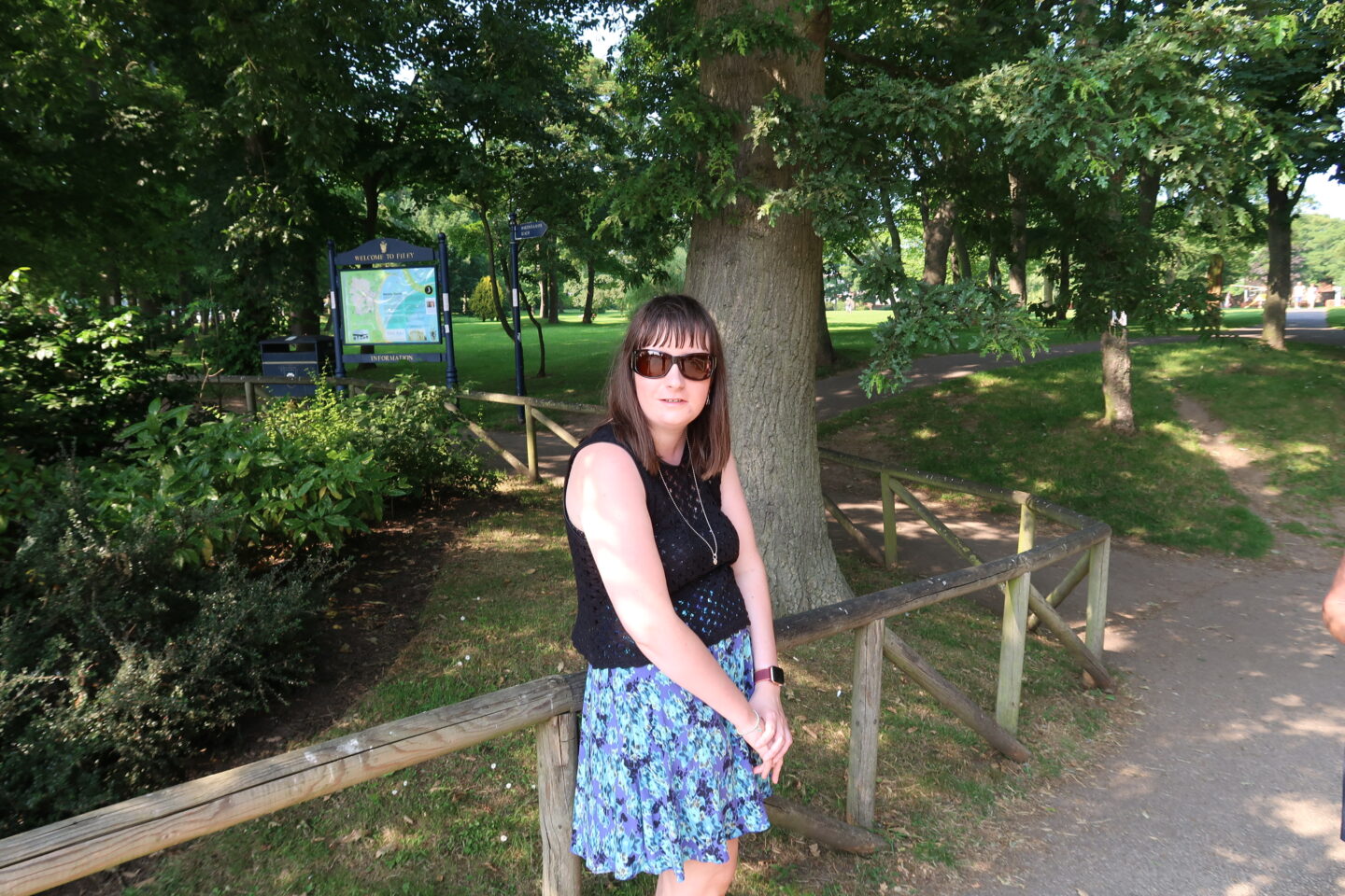 Holly leant against a wooden fence, she is looking directly at the camera. She's wearing a blue skirt, a black crocheted top, a necklace and sunglasses. There are green trees and grass in the background, the sun can be seen poking through trees in the distance