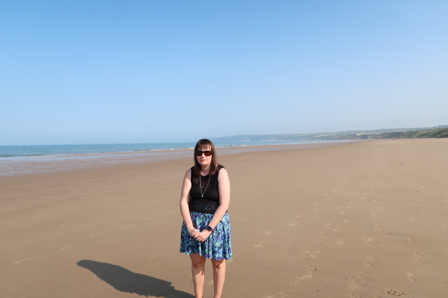 Holly stood on a beach with the sea in the background, she has her hands clasped in front of her and is wearing a blue skirt, a black crocheted top, a necklace and sunglasses
