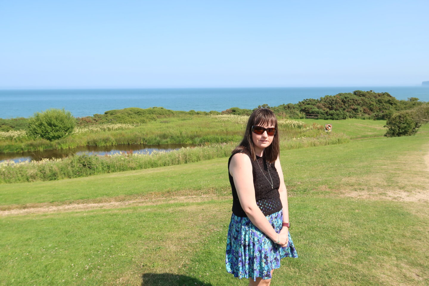 Holly stood sideways, she is wearing a blue skirt, a black crocheted top, a necklace and sunglasses. There is a grassy area and a lake behind her and the sea can be seen in the distance