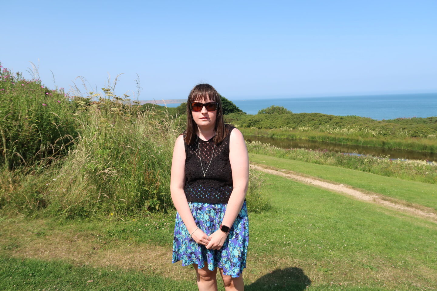 Holly stood with her hands clasped together in front of her, she is wearing a blue skirt, a black crocheted top, a necklace and sunglasses. A grassy area and a lake are behind her, the sea is in the distance