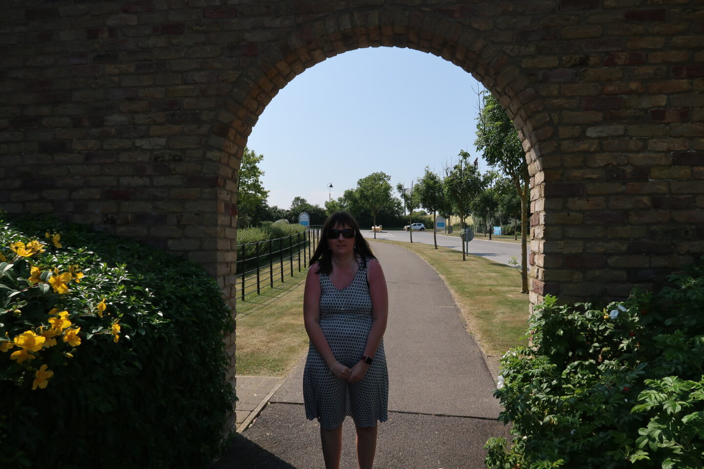 Holly stood in a brick archway which has flowers to the side, there is a line of trees behind her. She is wearing sunglasses and a white dress which has black detailing on it