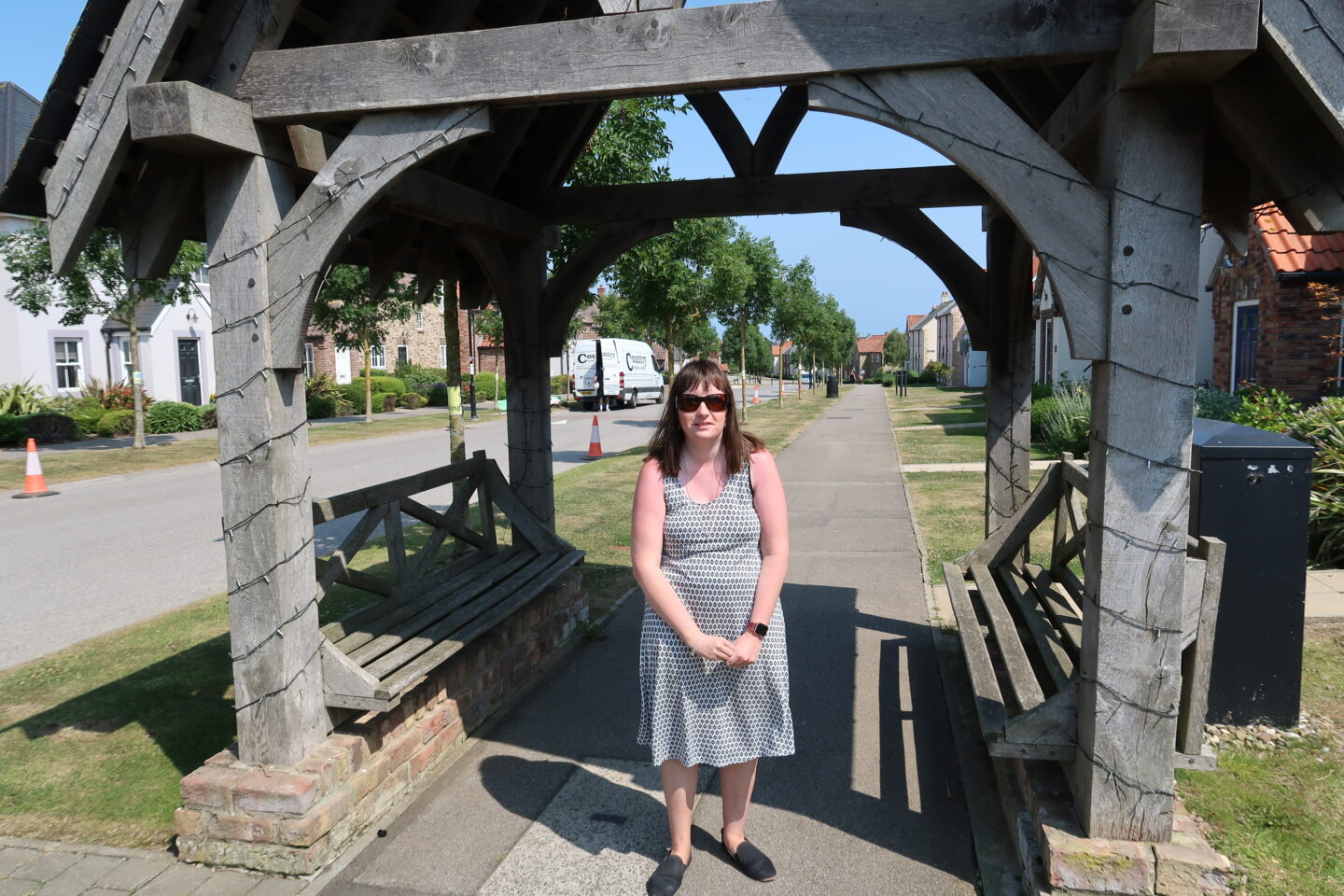 Holly stood in a wooden archway, buildings can be seen in the distance. She is wearing a white dress which has a black pattern on it, black shoes and sunglasses