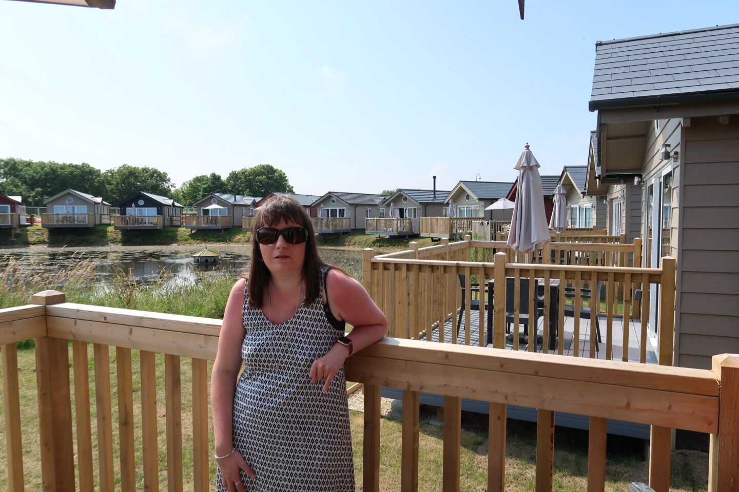 Holly stood on wooden decking which has a wooden fence surrounding it, she has one arm resting on the fence. She is wearing sunglasses and a white dress which has black detailing on it. There is a lake and log cabins in the background