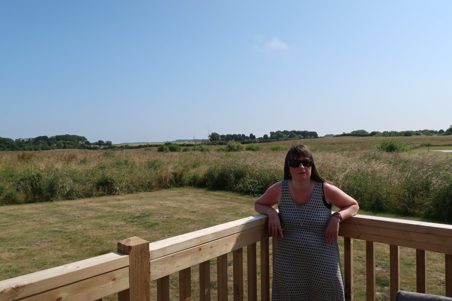 Holly stood on wooden decking with her elbows resting on a wooden fence, there are fields in the background. She is wearing sunglasses and a white dress which has a black pattern on it