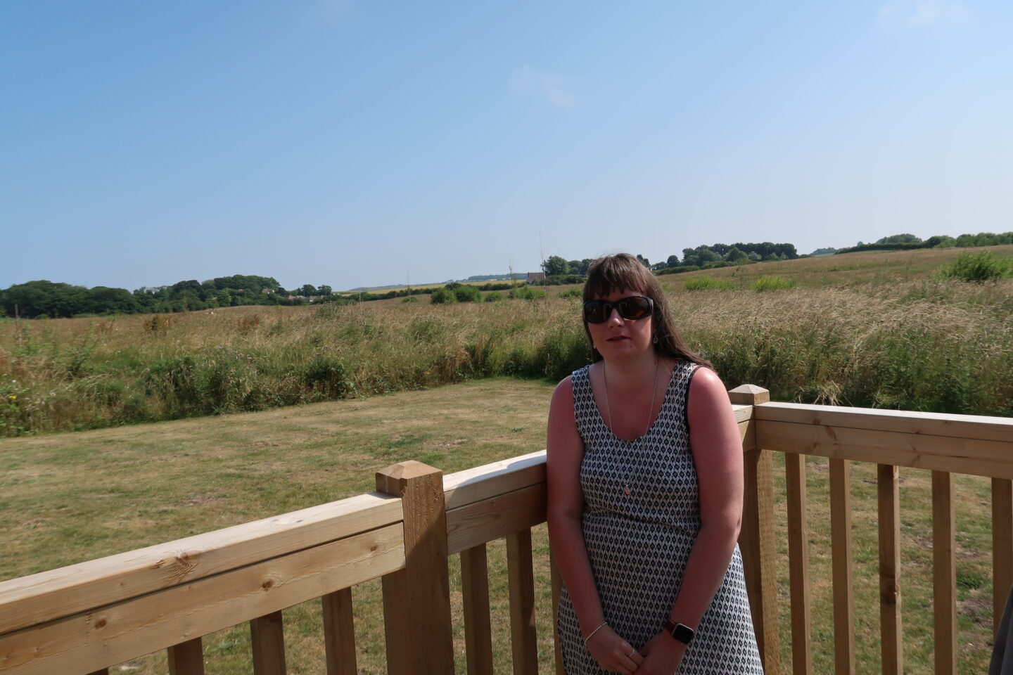 Holly stood on wooden decking which has a fence surrounding it, there are fields in the background. She is wearing sunglasses and a white dress which has a black pattern on it