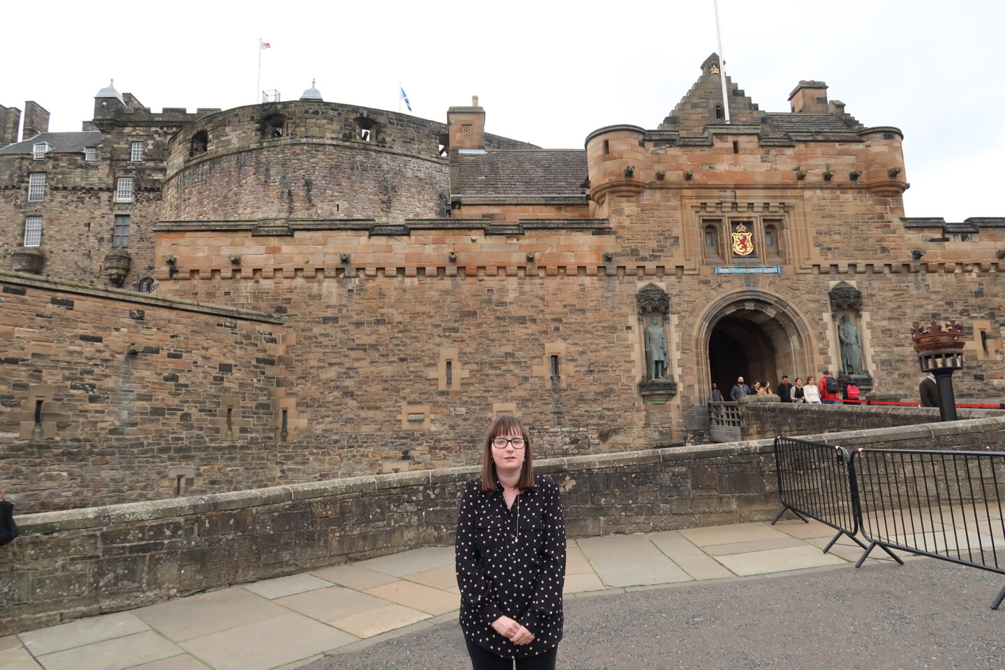 Holly stood in front of a stone wall, Edinburgh Castle can be seen in the background behind her. She's wearing a Black dotted blouse and black leggings