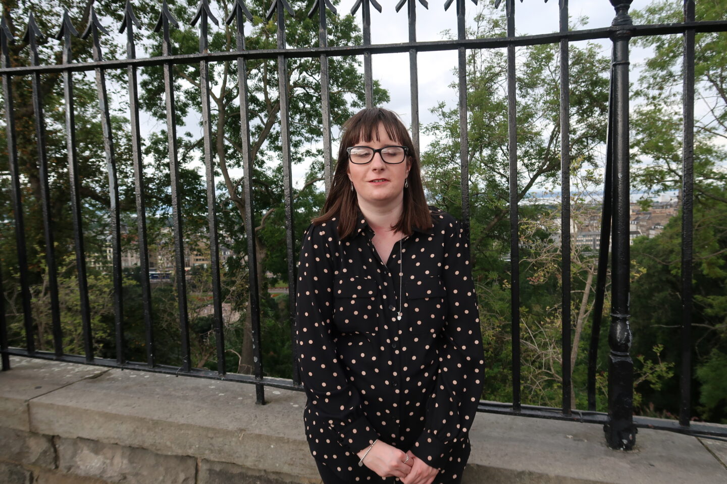 Holly stood in front of black metal railings, she is wearing a black dotted blouse. The city of Edinburgh can be seen in the distance behind a cluster of trees