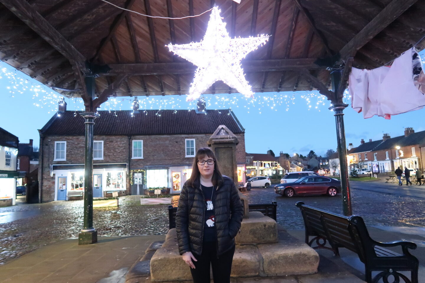 Holly stood in a town square with Christmas decorations all around, there is a star behind her. She is wearing a black jacket, navy Christmas jumper and black leggings