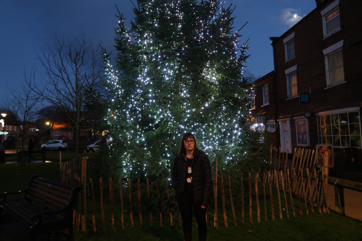 Holly stood in front of a green Christmas tree which has white lights on it, she is wearing a black jacket, a navy Christmas jumper which has polar bears on and black leggings