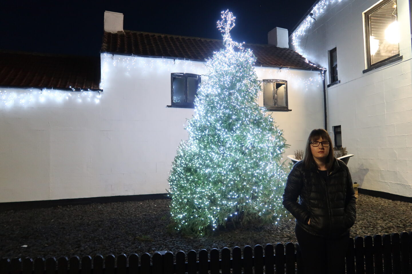 Holly stood in front of a green Christmas tree which has white lights on it, there is a white building in the background behind her. She is wearing a black jacket and black leggings and has her hands in her pockets