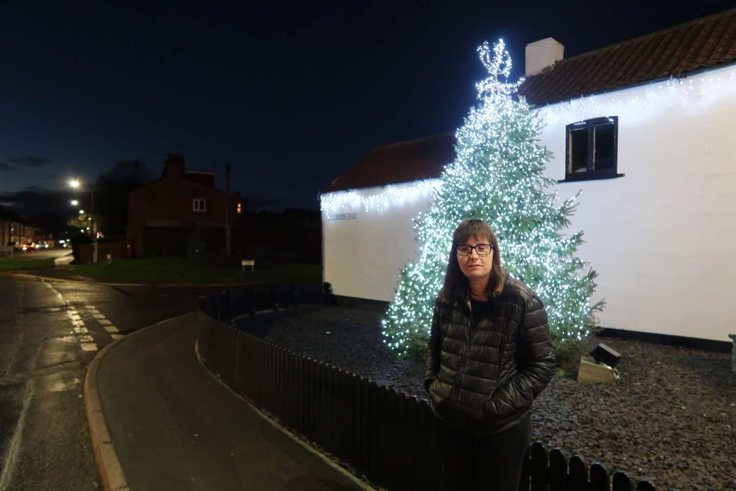 Holly stood in front of a green Christmas tree which has white lights on it, there is a white building in the background behind her. She is wearing a black jacket and black leggings and is stood to the side