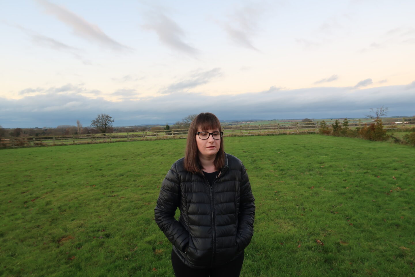 Holly stood with her hands in her pockets, she is wearing a black jacket and black leggings. There are fields and hills in the background