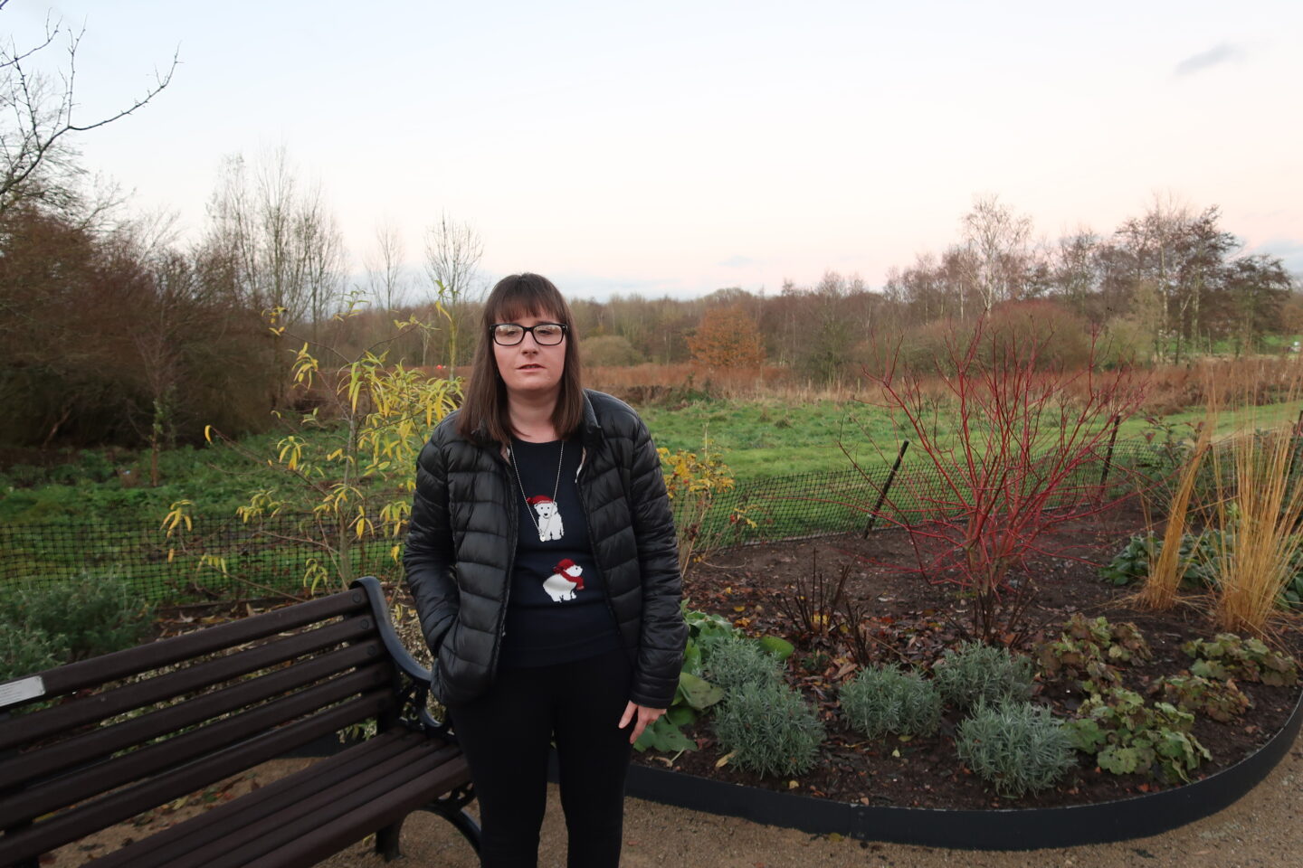 Holly stood with one hand in her pocket, she is wearing a black jacket, a navy Christmas jumper which has polar bears on it, there is multi-coloured shrubbery and bushes in the background behind her
