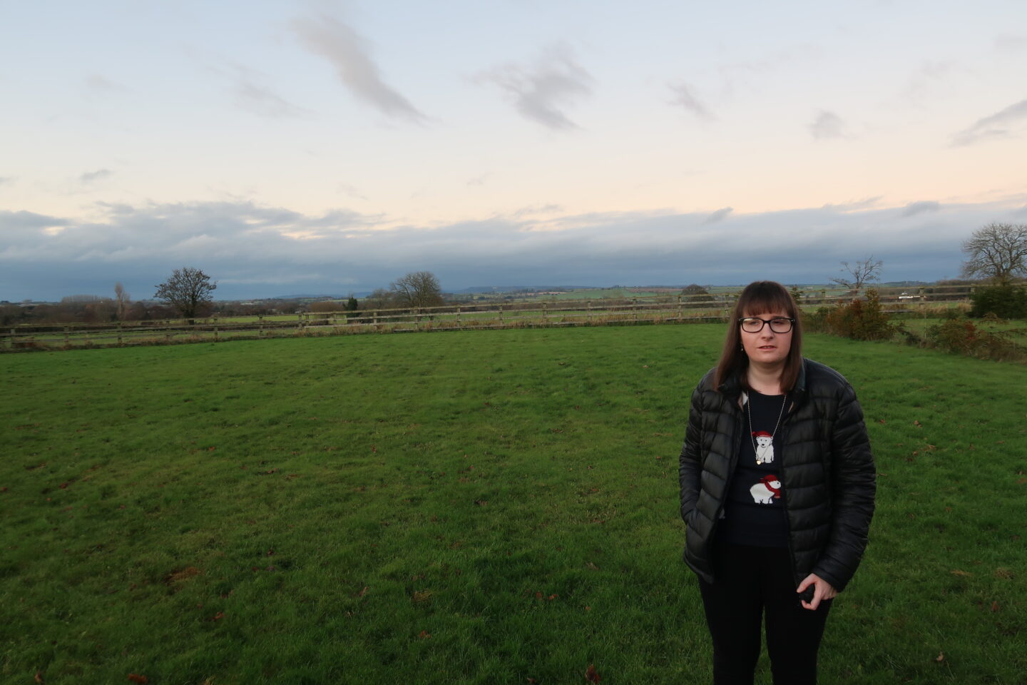 Holly stood with one hand in her pocket, she is wearing a Christmas jumper which has polar bears on it and black leggings. There are green fields and hills in the background