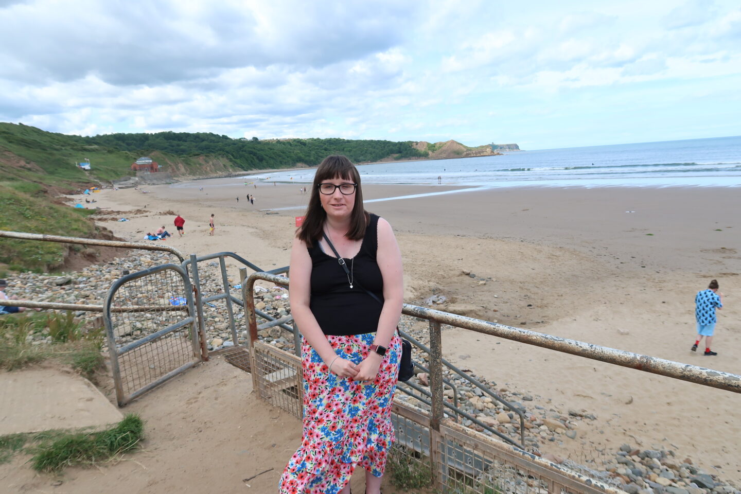 Holly stood beside some railings, the beach, see and cliffs are in the background. She is wearing a black top and a multicoloured flowery skirt