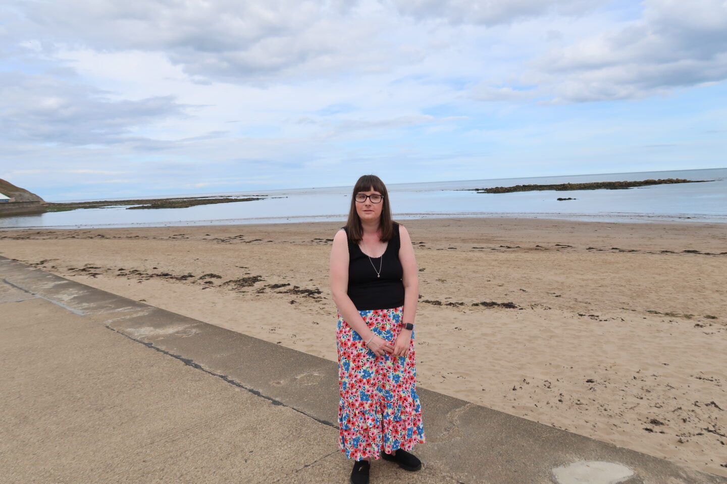 Holly stood on a stone path, the beach and sea are in the background. She is wearing a black top and a multicoloured flowery skirt