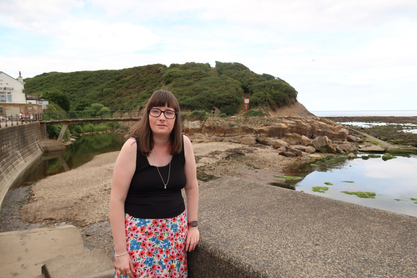 Holly stood with rocks behind her, she is wearing a black top and a long multicoloured flowery skirt