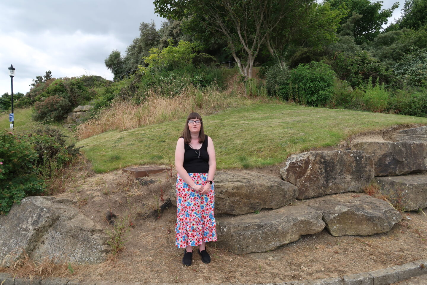 Holly stood with rocks behind her, there are trees and green grass in the background. She is wearing a black top and a long multicoloured flowery skirt