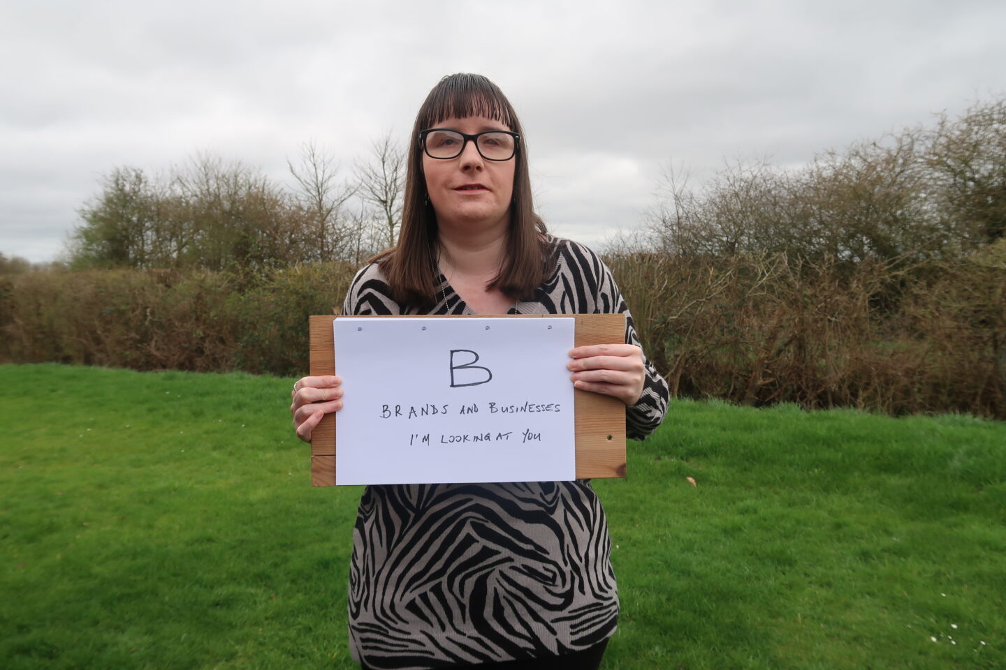 Holly stood on a grassy bank holding up a piece of paper on a wooden board which reads “B: Brands and businesses, I’m looking at you”. She’s wearing a grey and black patterned jumper and black leggings.