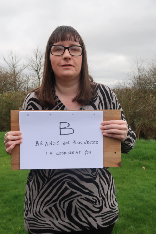 Holly stood on a grassy bank holding up a piece of paper on a wooden board which reads “B: Brands and businesses, I’m looking at you”. She’s wearing a grey and black patterned jumper and black leggings.