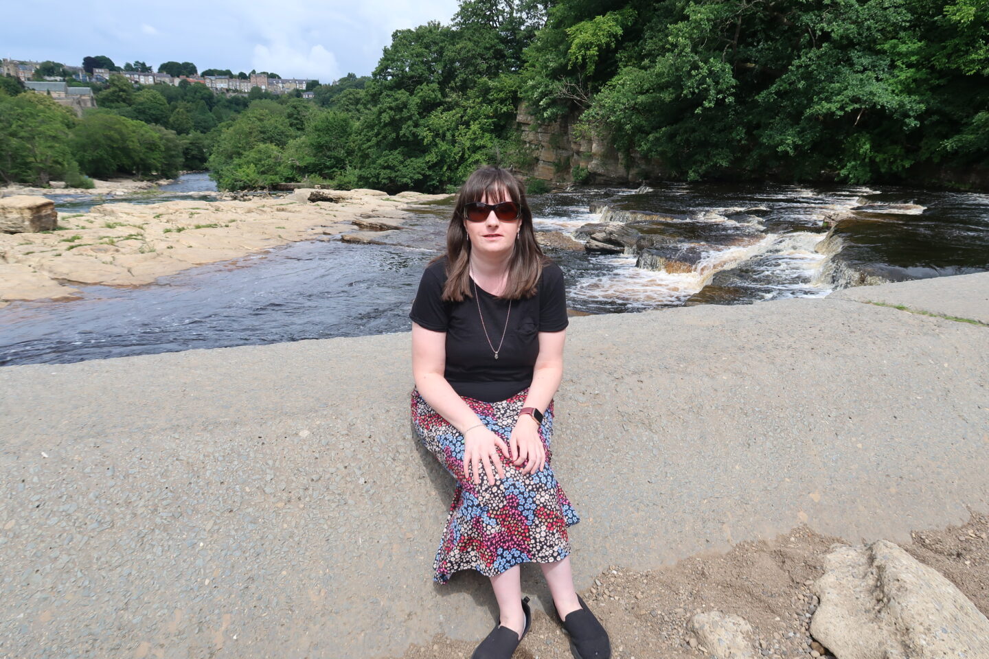 Holly sat on a stone wall, she's wearing a black t-shirt, a multicoloured skirt, black shoes and sunglasses. A waterfall can be seen protruding into a river in the background, and there are green trees in the distance.