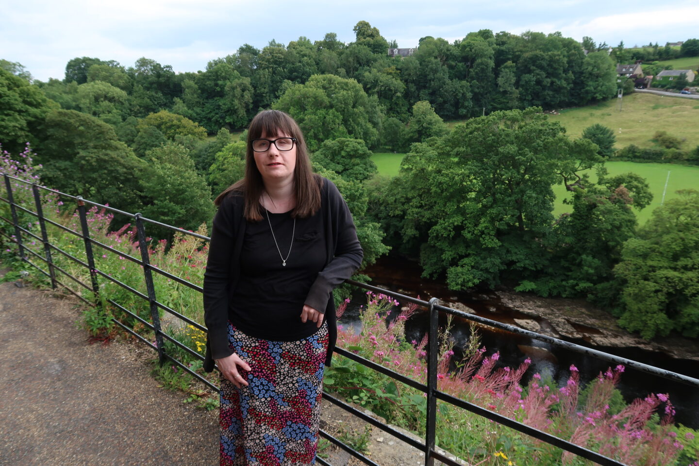 Holly stood with her arm on an iron railing, a cluster of green trees and a stream can be seen in the background. She's wearing a black cardigan, a black top and a multicoloured skirt.