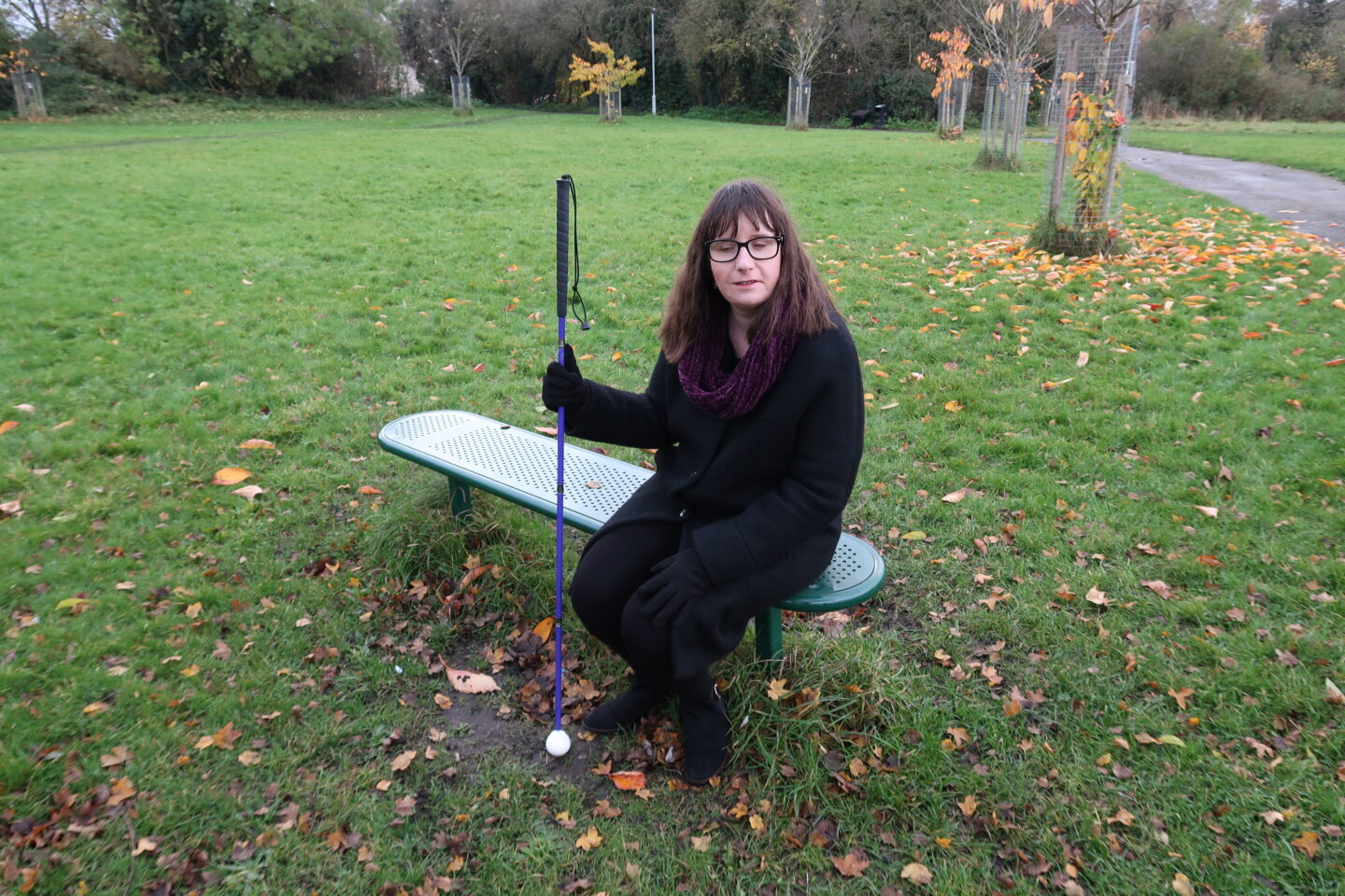 Holly sat on a bench holding her purple cane in her right hand. Fallen Autumn leaves can be seen on the ground. She's wearing a black coat, a burgundy scarf, black leggings and black boots.