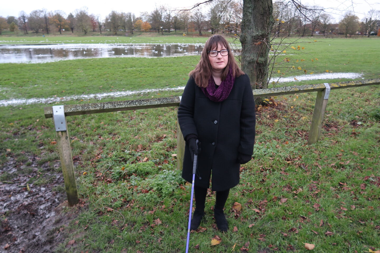 Holly stood in front of a wooden railing, a flooded field with a flock of seagulls can be seen in the distance behind her. She's wearing a black coat, a burgundy scarf, black leggings and black boots, and is holding her purple cane in her right hand.