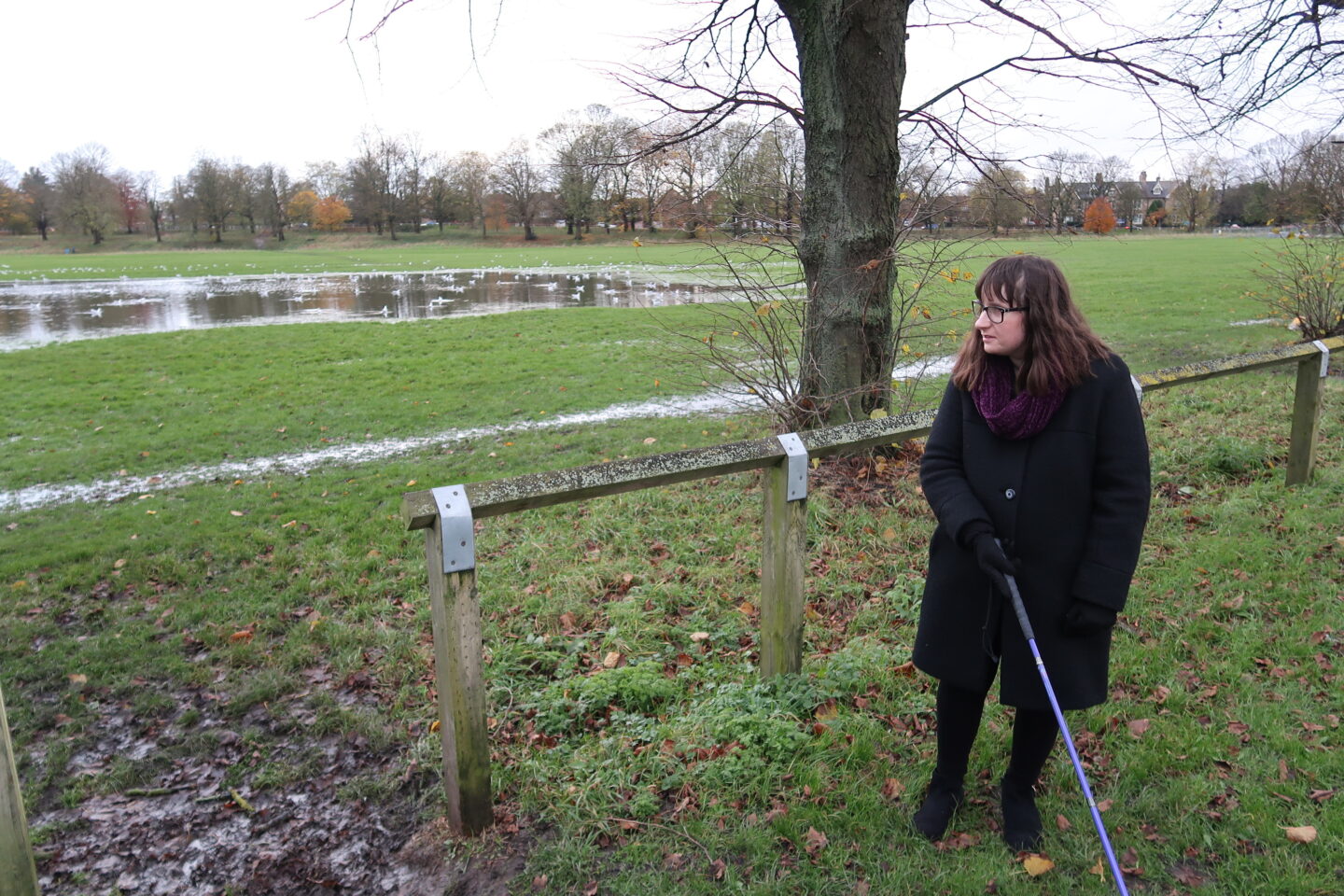 Holly stood in front of a wooden railing looking away from the camera with her long cane in her right hand. A flooded field with a flock of seagulls can be seen in the distance. She's wearing a black coat, a burgundy scarf, black leggings and black boots.