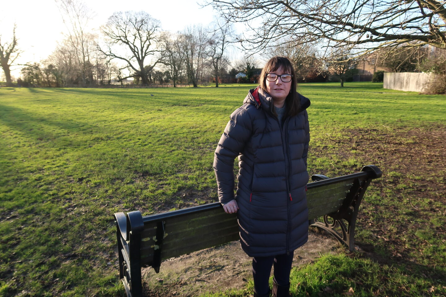 Holly leaning against the back of a bench with her hands placed on it. She's wearing a long dark blue coat, a light blue jumper, blue jeggings and black Ugg boots. There is a green grassy area and bare winter trees in the background.