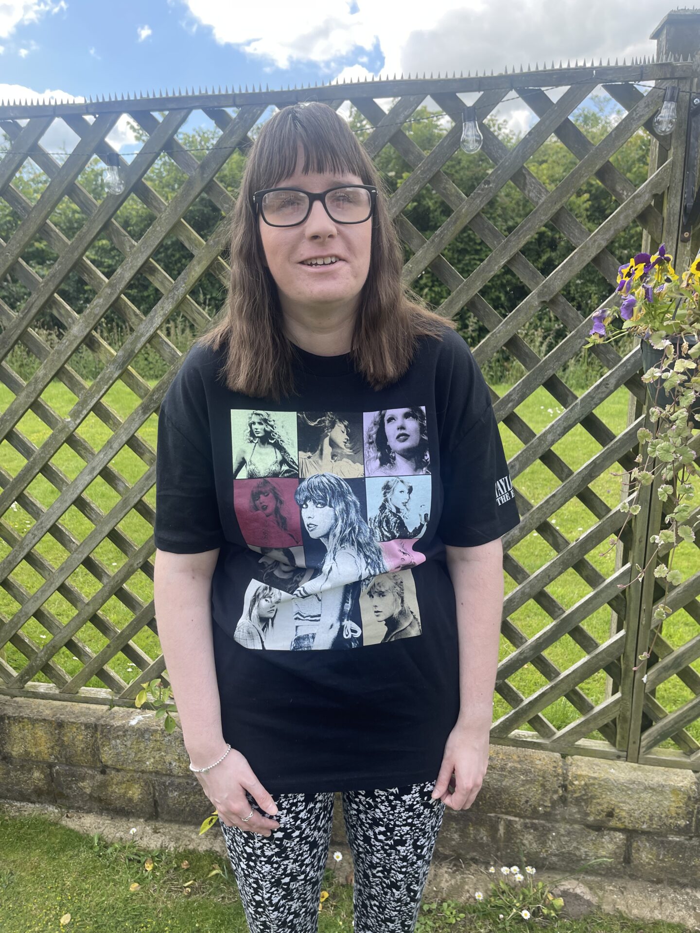 Holly stood in a garden with a fence and hedges behind her. She's wearing a black Taylor Swift t-shirt which features several images of Taylor Swift, and black and white flowery trousers.