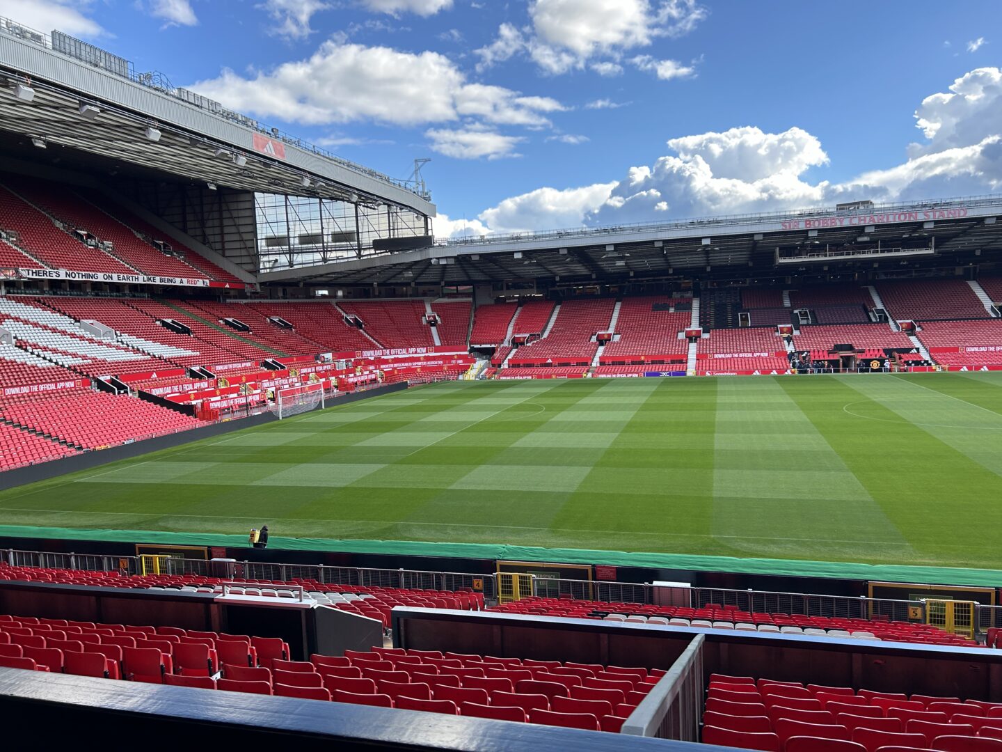 A view of an empty Old Trafford stadium with red seats in the stands, looking towards the Bobby Charlton stand. There is a banner that reads "There's nothing on Earth like being a red" along the upper part of the stands.